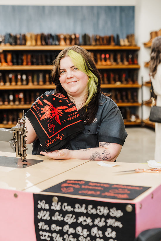 Kait is sitting at behind her embroidery machine inside a Boot Barn retail store, holding a black bandana with red screenprinting and 
