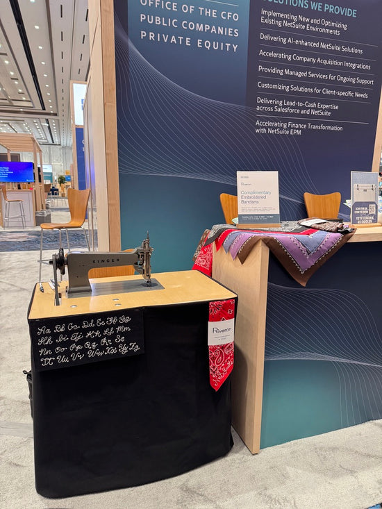 A vintage Singer embroidery machine displayed on a black draped table at a business expo booth, next to a wooden counter with a patterned textile draped over it, against a blue and white corporate backdrop. A small sign reading 
