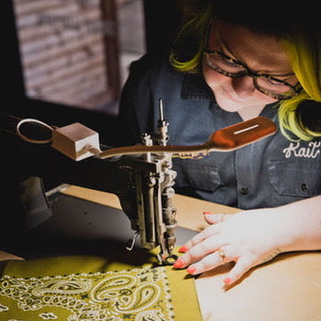 A person with glasses and bright green hair guides a bandana under an embroidery machine at a wedding welcome party.