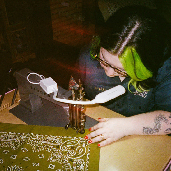 A person with bright green hair and glasses embroiders on an olive green paisley bandana with a white printed pattern using a vintage Singer sewing machine under a white lamp.