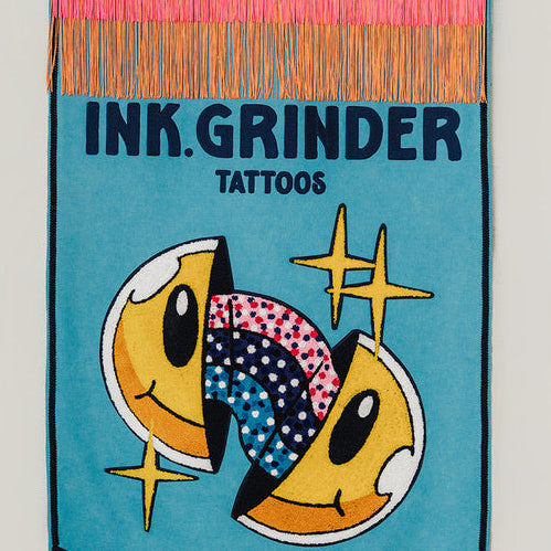 A vertical fabric banner with a teal background, pink and orange fringe, and the text 'INK GRINDER TATTOOS' above two stylized yellow smiley faces with colorful patterned eyes and gold star accents, hanging against a light gray wall.
