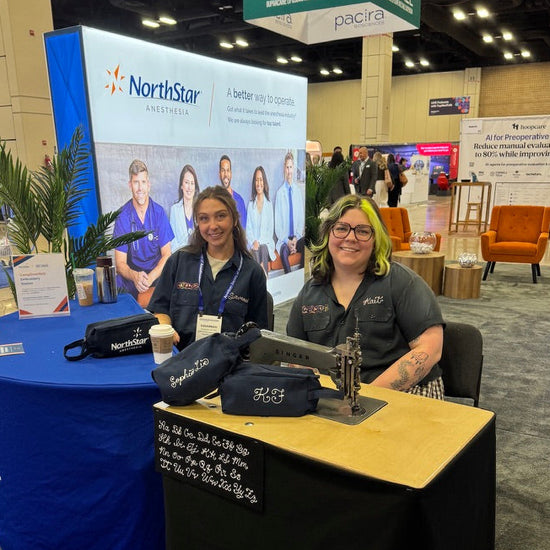 Kait and embroidery assistant Savannah are seated behind their client NorthStar Anesthesia's booth at a trade show or convention, with NorthStar branding visible on a large backdrop and a conference sign overhead. The setup includes a blue draped table, a black table with a light wood top, embroidery machines, and embroidered black bags.
