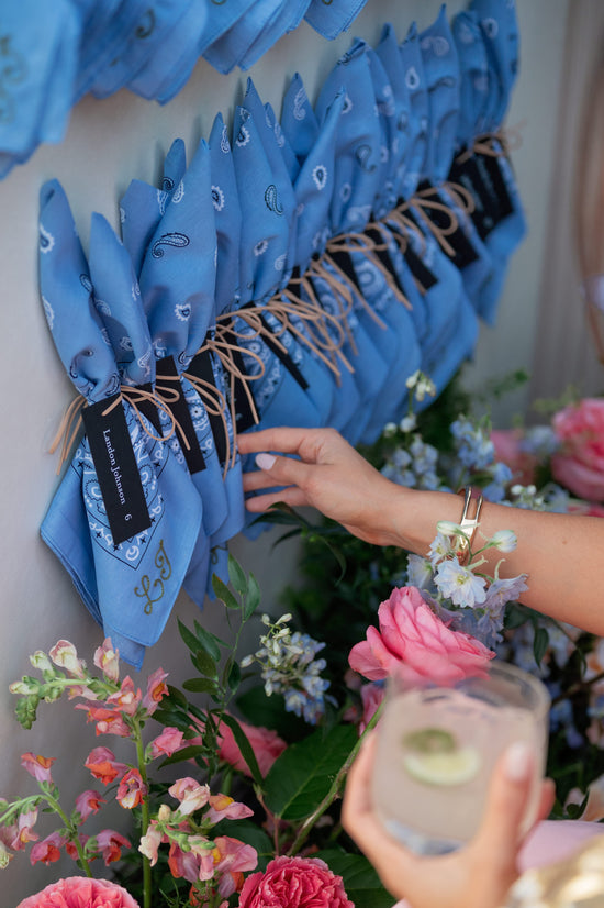 Hand holding a glass with flowers and monogrammed blue bandanas hanging on a wall.